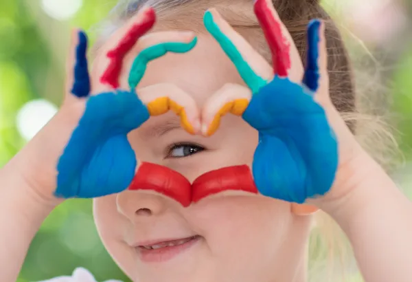 Kind formt mit bunt bemalten Händen ein Herz vor seinem Gesicht