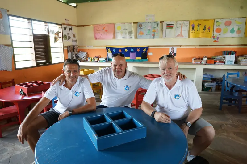 Drei Besucher sitzen an einem blauen Kindertisch in einem neu eingerichteten Klassenraum
